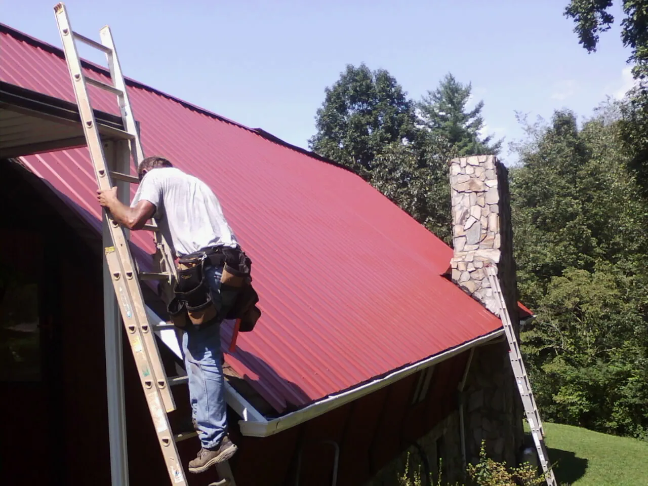 Roofer on a ladder beside a red metal roof and stone chimney — Soffit & Fascia Repair by Roofing Brothers in Siloam